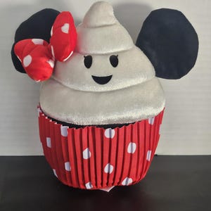 May include: A plush cupcake toy with a white frosting top and a red polka-dot cupcake liner. The cupcake has black ears and a red bow with white polka dots. The frosting has a smiling face.