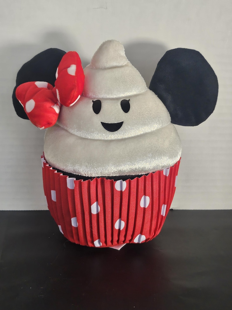May include: A plush cupcake toy with a white frosting top and a red polka-dot cupcake liner. The cupcake has black ears and a red bow with white polka dots. The frosting has a smiling face.