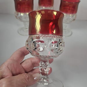 May include: Set of four clear glass goblets with a red iridescent rim. The goblets have a decorative, textured design on the bowl and a clear stem and base. The glasses are suitable for serving beverages.