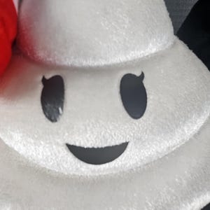 May include: A plush white character with a smiling face, featuring black eyes and a curved mouth. The character has a soft, textured appearance, suggesting a stuffed toy or pillow. The design is simple and cartoonish.