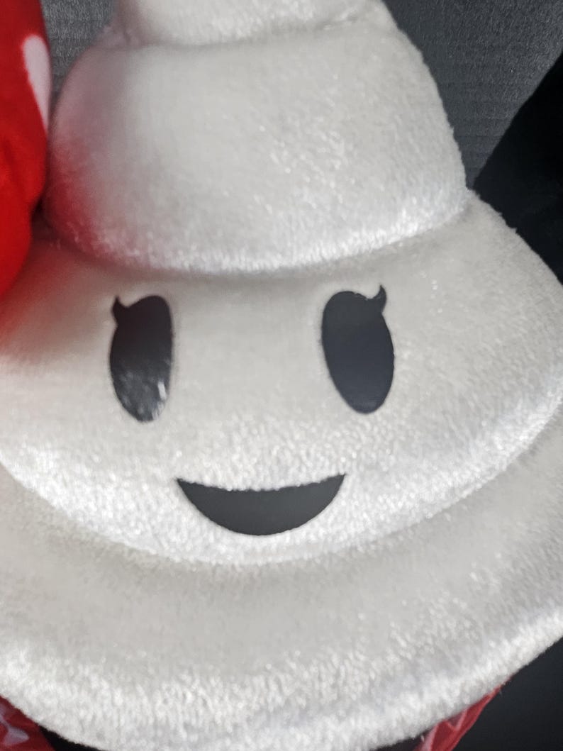 May include: A plush white character with a smiling face, featuring black eyes and a curved mouth. The character has a soft, textured appearance, suggesting a stuffed toy or pillow. The design is simple and cartoonish.