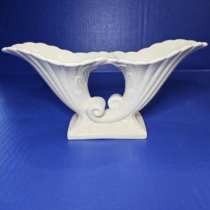 May include: White ceramic vase with a unique, sculpted design. The vase has a wide, open top and a narrow base. It features two curved, trumpet-like openings on either side.