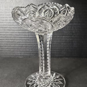 May include: A clear glass pedestal bowl with a scalloped edge and intricate cut glass design. The bowl sits on a tall, slender stem with a starburst-shaped base.