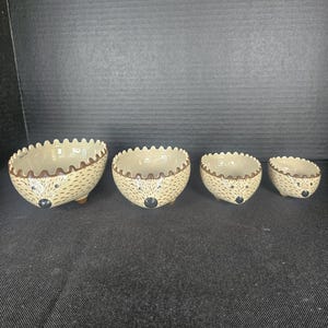 May include: A set of four graduated ceramic bowls shaped like hedgehogs. Each bowl features a cream-colored interior, a brown scalloped rim, and a tan exterior with brown speckles, a painted black nose, and small brown feet on the base.