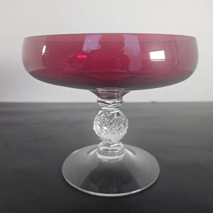 May include: A vintage coupe glass with a deep red bowl and a clear, decorative stem. The stem features a textured, spherical design, and the base is a clear, circular foot. The glass is designed for serving champagne or cocktails.