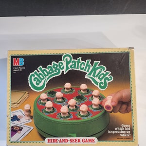 May include: A green and red game board with the words "Cabbage Patch Kids" and "Hide-and-Seek Game". The board has a circular opening with 12 small holes, each with a small plastic figure of a Cabbage Patch Kid peeking out. A hand is holding a figure of a Cabbage Patch Kid.
