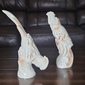 May include: Two ceramic parrot figurines. One parrot is perched upright, the other is in a more abstract pose. Both are white with peach-colored accents. The figurines are on a dark wooden surface, with a dark brown leather couch in the background.