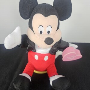 May include: A plush Mickey Mouse toy featuring large black ears, a tan face, and a black nose. The character wears a black jacket over a white shirt, red trousers with two white buttons, and black shoes. It holds a pink heart with the text BE MINE.