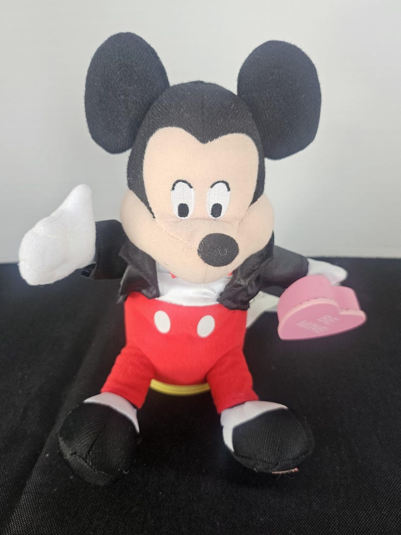 May include: A plush Mickey Mouse toy featuring large black ears, a tan face, and a black nose. The character wears a black jacket over a white shirt, red trousers with two white buttons, and black shoes. It holds a pink heart with the text BE MINE.