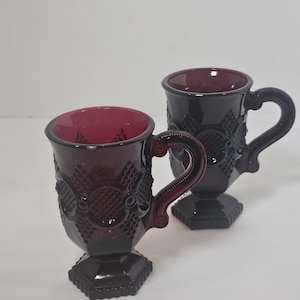 May include: Two ruby red glass mugs with ornate, raised patterns and pedestal bases. The mugs have curved handles and a deep red interior. The design features geometric shapes and floral-like details, creating a vintage aesthetic.