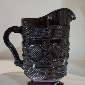 May include: A dark purple glass pitcher with a decorative pattern. The pitcher has a wide mouth and a handle.
