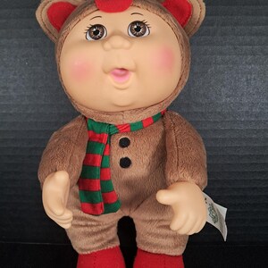 May include: A Cabbage Patch Kids doll dressed in a brown plush reindeer costume with a hood featuring ears and antlers. The doll has a red nose, pink cheeks, and brown eyes. It wears a red and green striped scarf and has red feet.