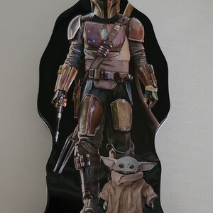 Puede incluir: Una fiambrera de metal negro con un personaje mandaloriano de la franquicia Star Wars. El Mandaloriano lleva un casco y una armadura y sostiene un arma. Un peque&ntilde;o personaje alien&iacute;gena verde est&aacute; de pie frente al Mandaloriano.