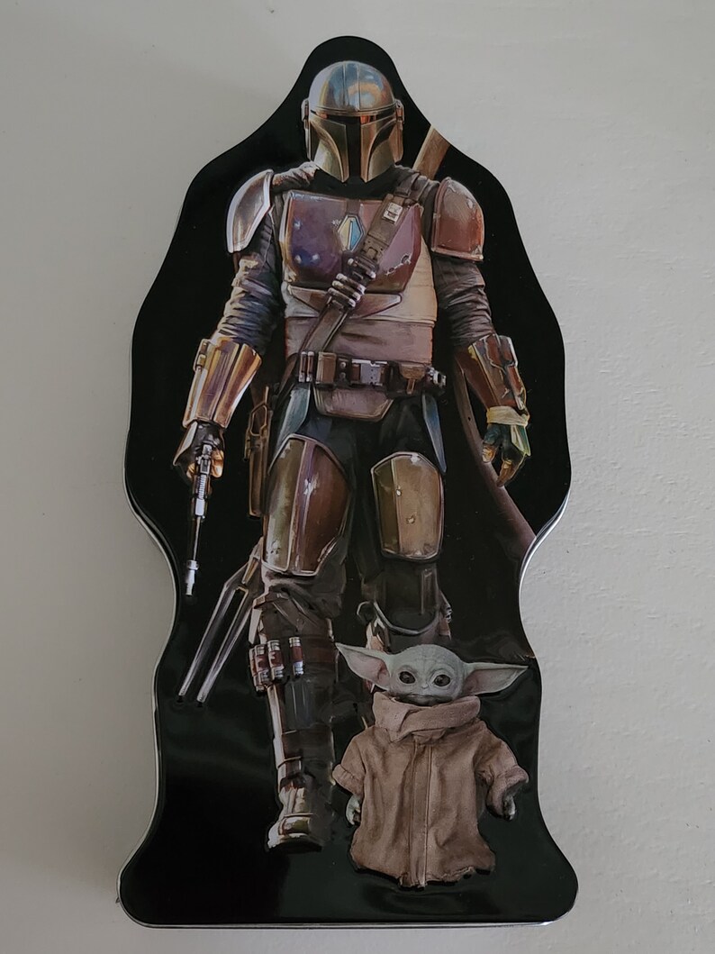 Puede incluir: Una fiambrera de metal negro con un personaje mandaloriano de la franquicia Star Wars. El Mandaloriano lleva un casco y una armadura y sostiene un arma. Un peque&ntilde;o personaje alien&iacute;gena verde est&aacute; de pie frente al Mandaloriano.