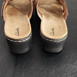 May include: A pair of brown leather slide sandals with a woven strap design across the top. The footbed is tan with the Clarks brand name printed in black. The sandals feature a black wedge heel with visible white stitching along the base.