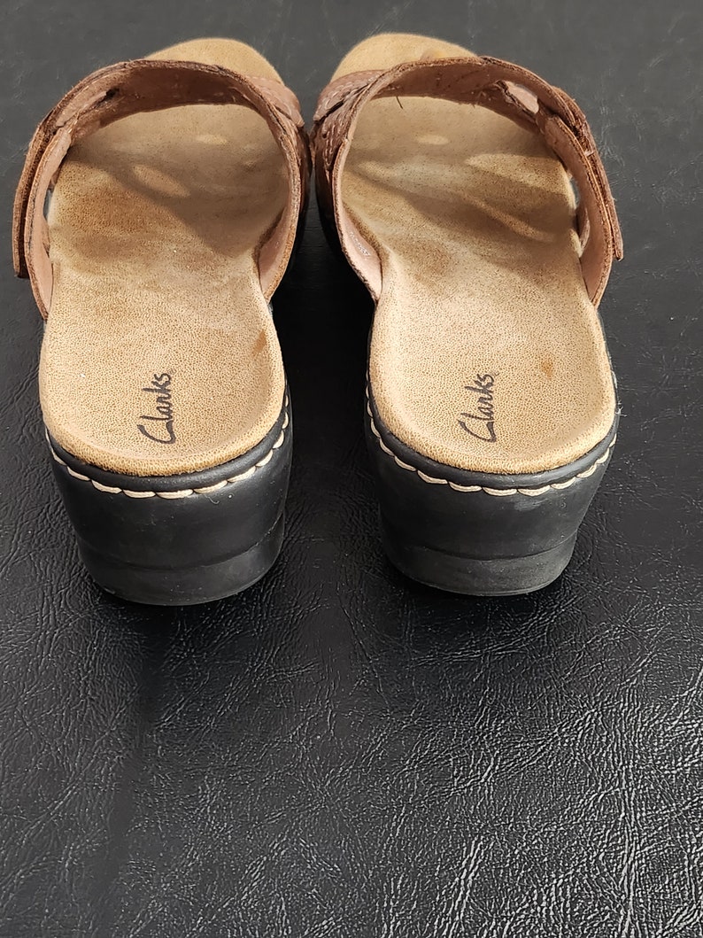 May include: A pair of brown leather slide sandals with a woven strap design across the top. The footbed is tan with the Clarks brand name printed in black. The sandals feature a black wedge heel with visible white stitching along the base.
