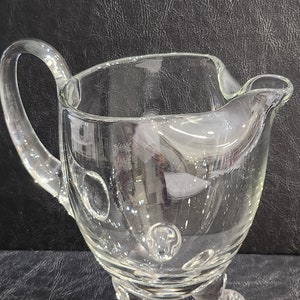 May include: A clear glass pitcher with a rounded body and a handle. The pitcher has three legs and a textured surface with small bubbles.