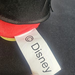 May include: A plush toy featuring a black fabric upper section and a bright red fabric lower section. A white fabric tag is attached to the side, clearly displaying the copyright symbol followed by the text Disney in black capital and lowercase letters.