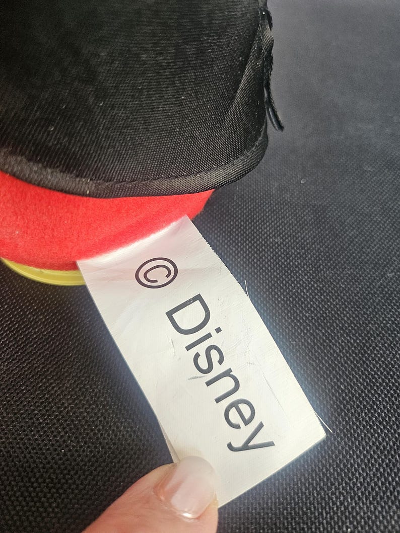 May include: A plush toy featuring a black fabric upper section and a bright red fabric lower section. A white fabric tag is attached to the side, clearly displaying the copyright symbol followed by the text Disney in black capital and lowercase letters.