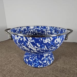 May include: A round colander featuring a blue and white marbled enamel finish. The bowl has a wide rim, two curved metal side handles, and a circular pedestal base. Multiple drainage holes are evenly spaced around the sides of the basin.