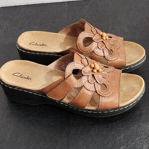 May include: A pair of brown leather slide sandals with a low wedge heel and a black sole. The upper features a decorative cutout flower design with small wooden beads in the center. The Clarks brand name is printed on the tan footbed of each sandal.