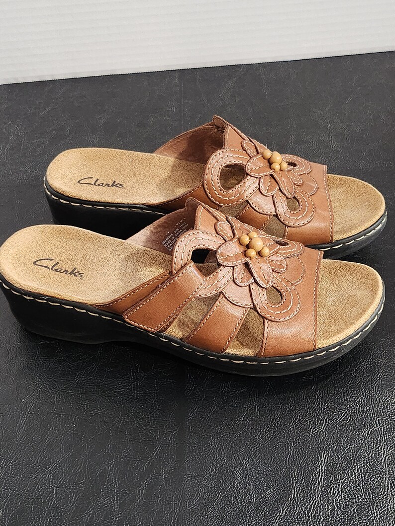 May include: A pair of brown leather slide sandals with a low wedge heel and a black sole. The upper features a decorative cutout flower design with small wooden beads in the center. The Clarks brand name is printed on the tan footbed of each sandal.