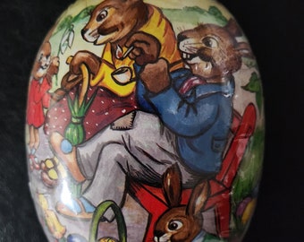 May include: A vintage Easter egg-shaped tin with a colorful illustration of four rabbits. The rabbits are sitting on a red bench and holding Easter eggs. The tin is decorated with a green basket filled with Easter eggs.