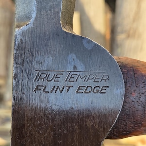 May include: Close-up of a vintage axe head with the text "TRUE TEMPER FLINT EDGE" etched into the metal. The axe head is a dark, weathered steel color with a wooden handle. The background is blurred.