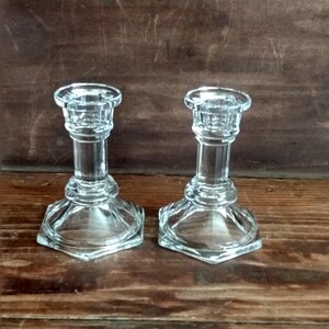 May include: Two clear glass candle holders. Each has a hexagonal base, a cylindrical stem, and a circular top designed to hold a candle. The candle holders are displayed on a wooden surface.