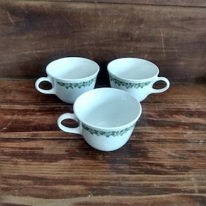 May include: Three white ceramic teacups with a green floral pattern around the rim. Each cup has a curved handle and sits on a dark wooden surface. The cups are empty and ready for use.