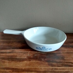 May include: A white, oval-shaped serving dish with a handle. The dish has a light blue floral pattern around the rim. The dish is sitting on a wooden surface.