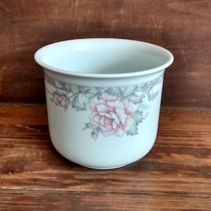 May include: A white ceramic planter with a rounded body and a slightly flared rim. The planter is decorated with a floral pattern featuring pink roses and gray leaves. The background is a wooden surface.