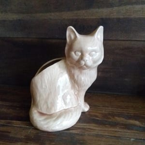 May include: A light pink ceramic cat figurine with a hollowed-out back, likely a planter or vase. The cat is in a seated position with its tail curled around. The figurine has a glossy finish and is set against a dark wood background.