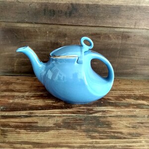 May include: A light blue ceramic teapot with a rounded body, a curved spout, and a loop handle. The lid has a small ring handle and a gold trim. The teapot is set against a wooden background.