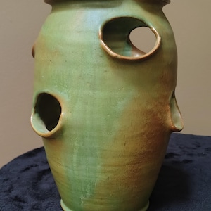 May include: A cylindrical ceramic planter with a green and brown glazed finish. The vessel features a wide, flared rim and multiple circular openings spaced around the body, designed for planting. The surface shows variations in color and texture.