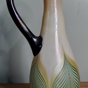 Puede incluir: Una jarra decorativa de vidrio con un asa negra. La jarra tiene un cuerpo color crema con patrones verdes y amarillos en forma de hojas. La base es verde y el asa es curva.