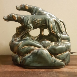 Op de afbeelding: Een keramische lampenvoet met twee gestileerde honden die op een rotsachtige groene en gouden basis staan.