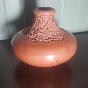 Puede incluir: Un jarrón de terracota con un diseño tallado alrededor de la parte superior y un acabado liso ligeramente brillante.