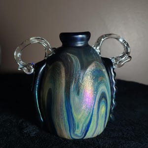 Puede incluir: Un jarrón de vidrio artesanal con un patrón arremolinado de azules, verdes y tonos iridiscentes. El jarrón presenta dos asas claras y curvas y un borde azul oscuro. Es una pieza de arte decorativa.