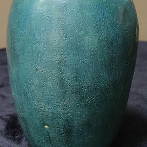 Op de afbeelding: Een blauwgroene keramische vaas met een ronde, bolvormige vorm en een smalle opening. Het oppervlak heeft een getextureerde, matte afwerking met zichtbare putjes en fijne craquelépatronen over de gehele buitenkant, met subtiele kleurvariaties.