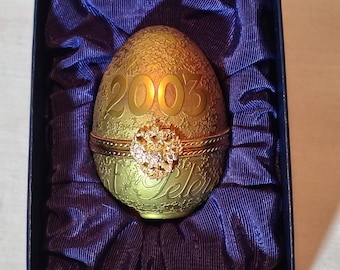 May include: A gold egg-shaped ornament with the year 2003 embossed on it. The ornament has a jeweled heart design and is in a blue velvet box.
