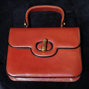 Op de afbeelding: Een vintage, roodbruine handtas met een handvat aan de bovenkant en een rechthoekige vorm. De tas heeft een goudkleurige sluiting en zwarte rand. De handgreep is ook roodbruin. De tas staat op een zwart oppervlak.