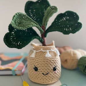 Puede incluir: Una planta de Fiddle Leaf Fig hecha a mano con un rostro sonriente en la maceta. La planta tiene hojas grandes de color verde oscuro y un tallo marrón. La maceta beige tiene borlas blancas y ojos y boca bordados en negro.