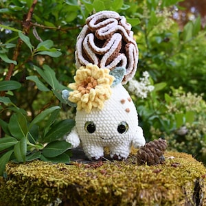 May include: A white crocheted creature with large black eyes and a small smile, featuring a brown and white textured mushroom cap on its head and a yellow flower accent. The figure has small arms and legs, with light brown spots on its side.