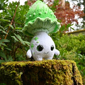 Peut inclure: Une figurine en crochet représentant un corps blanc arrondi avec de petits bras et jambes, orné de broderies en forme d'étoiles vertes. La figurine est surmontée d'un grand chapeau de champignon vert clair texturé à bord volanté.