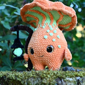May include: A plush crochet mushroom creature with a large orange cap, a light green underside, and a textured orange body featuring small light green spots. The figure has large black eyes, a small stitched mouth, and holds a black lantern.