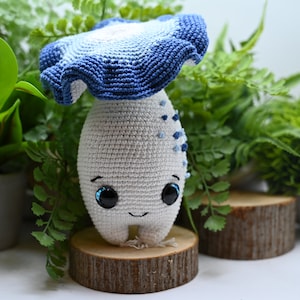 May include: A crocheted mushroom toy with a blue and white cap and a white body. The mushroom has black eyes and a smiling mouth. The toy is sitting on a wooden base.