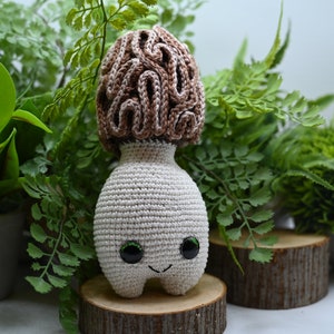May include: A crocheted mushroom toy with a brown and white cap and a white body. The toy has green eyes and a smiling mouth. The toy is sitting on a wooden base.