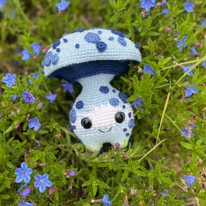 Puede incluir: Un juguete de peluche de hongo azul tejido a crochet con detalles blancos y ojos grandes. El juguete está sentado en una cama de flores azules.