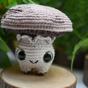 May include: A crocheted mushroom toy with a brown cap and a white body. The toy has large, black eyes and a smiling mouth.